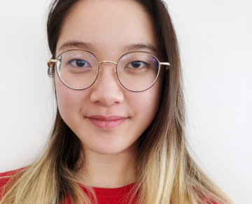 A photograph of Mikaela. Mikaela has blonde hair, is wearing glasses and a red jumper, and is looking towards the camera.