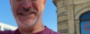 A photograph of Stuart. Stuart is looking towards the camera and smiling outside in front of a building, against a bright blue sky.