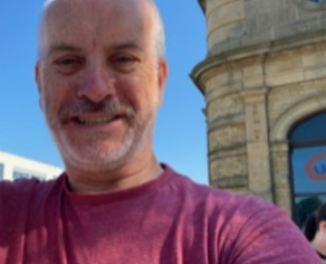 A photograph of Stuart. Stuart is looking towards the camera and smiling outside in front of a building, against a bright blue sky.