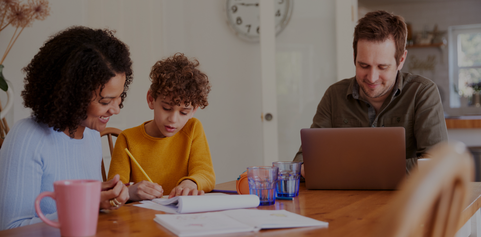 A photograph showing a middle-aged man, his partner and their son at the kitchen table. The man is smiling whilst working on his laptop. The partner and son are writing on paper opposite. Representing an IET member renewing their IET membership online.