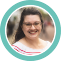The photo shows Catherine Drake, Communications and Digital Event Officer. She has long brown hair and green eyes, and wears transparent pink-framed glasses. She is smiling at the camera.