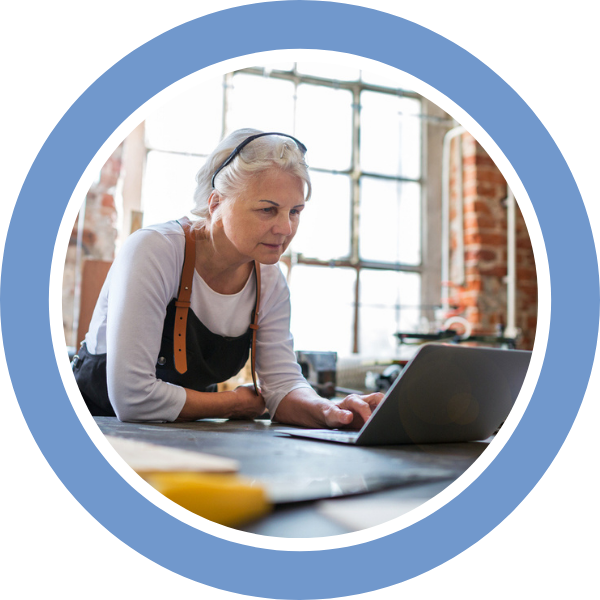 Woman in overalls and her glasses on her head leans over a workbench to browse on her laptop.