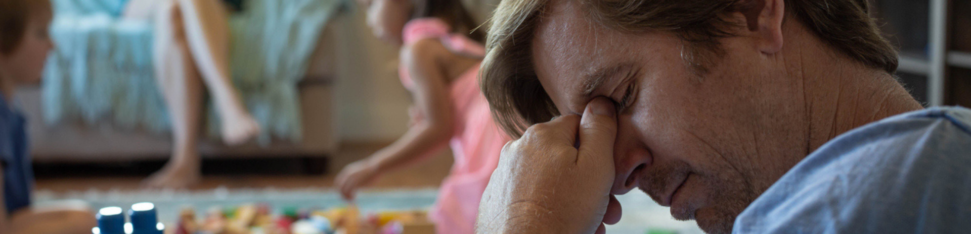 A photograph showing a middle-aged man looking concerned with his head in his hand, whilst his young family play together in the background - representing an engineering family that is facing crisis and needs support